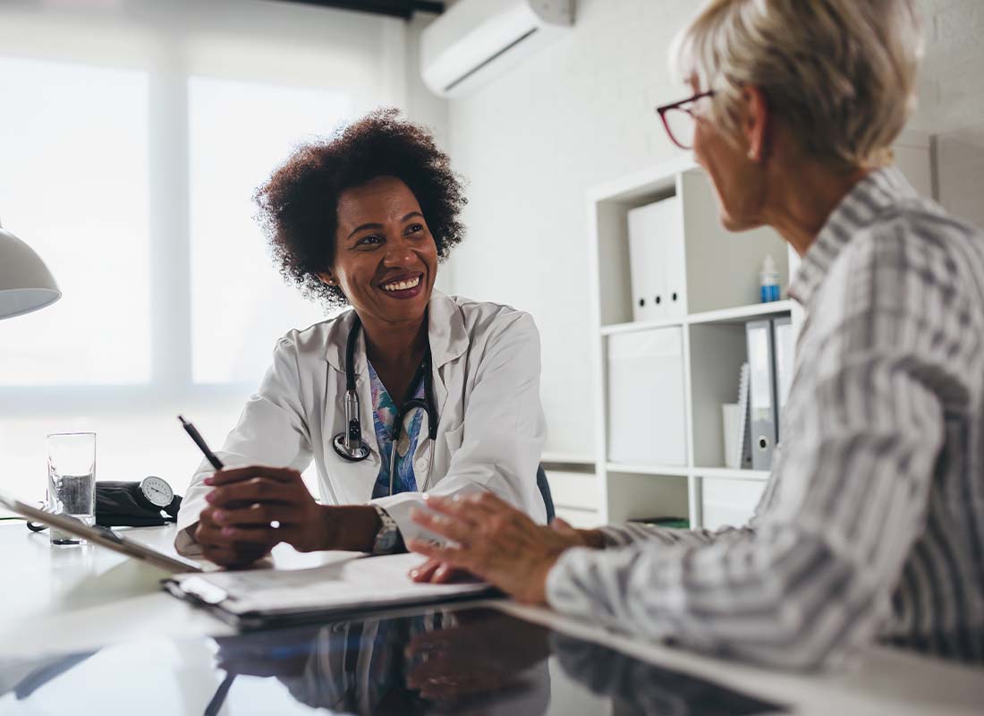 Medical Advantage Plans - Smiling Young Doctor Sits at Her Office Desk and Talks with an Elderly Female Patient about Her Healthcare Options