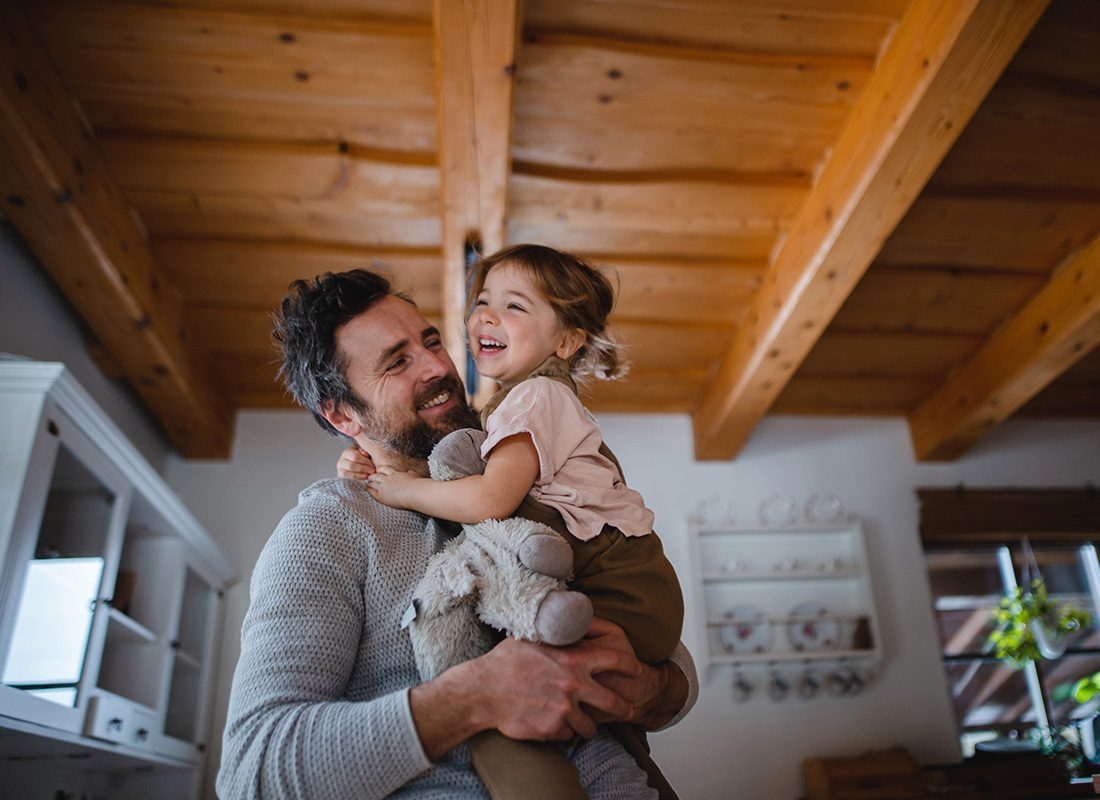 About Our Agency - Closeup Portrait of a Cheerful Father Holding Up his Daughter with a Stuffed Animal in the Kitchen with a Wooden Beam Ceiling