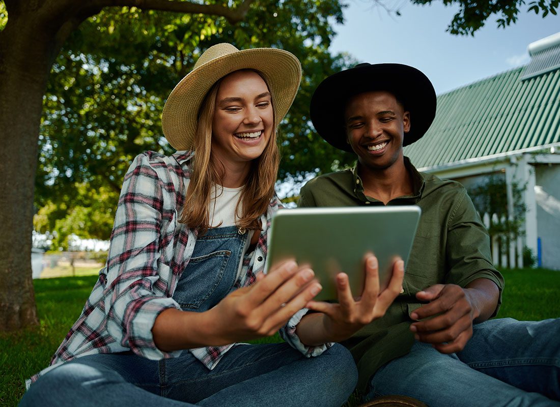 Blog - Closeup Portrait of Young Male and Female Farmers Sitting Together on the Green Grass Next to a Tree While Using a Tablet Together