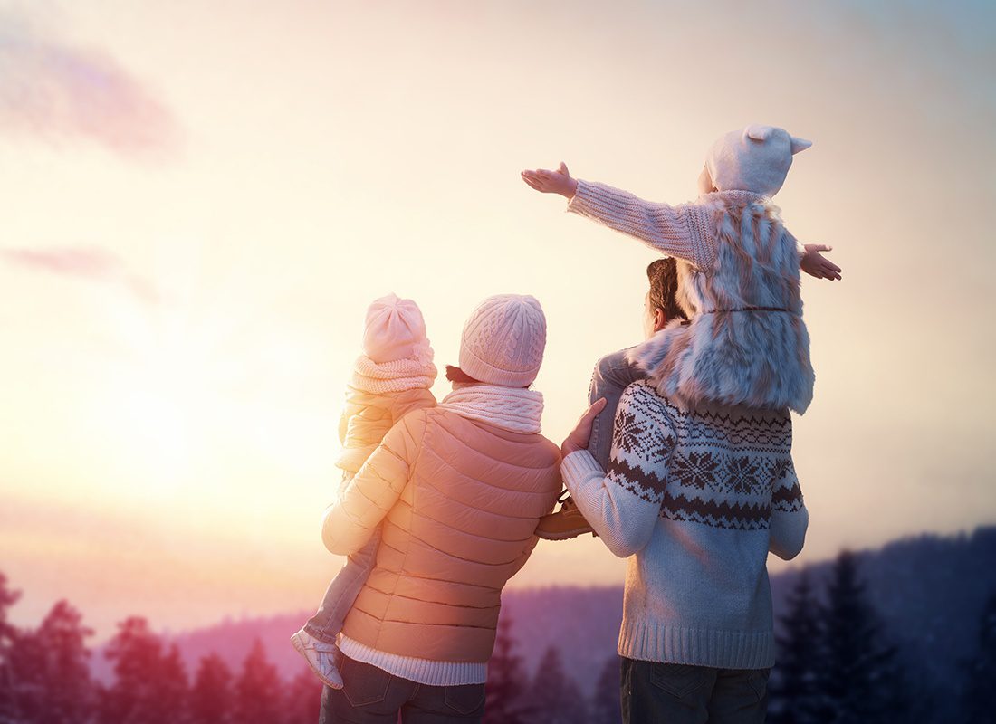 Insurance Solutions - Rear View of a Family with Two Young Kids Wearing Hats and Coats Standing in the Park During the Winter Watching the Sunset Over the Mountains and Trees
